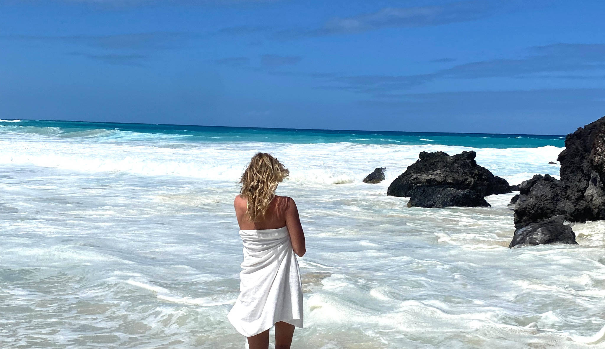 Woman in a white dress standing on a beach with blue sky and ocean.